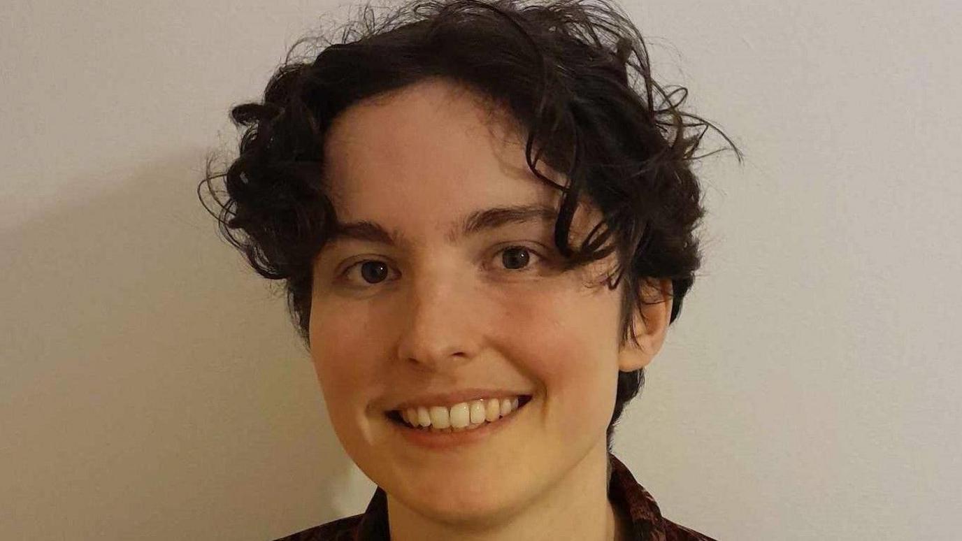 A person smiling at the camera with a blank wall in the background