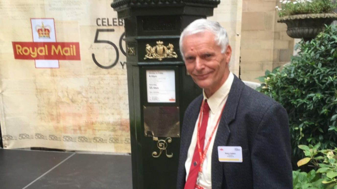 Postman Robert Hudson stands in front of a special postbox to mark 50 years of the royal mail. Robert is wearing a suit with a red tie.