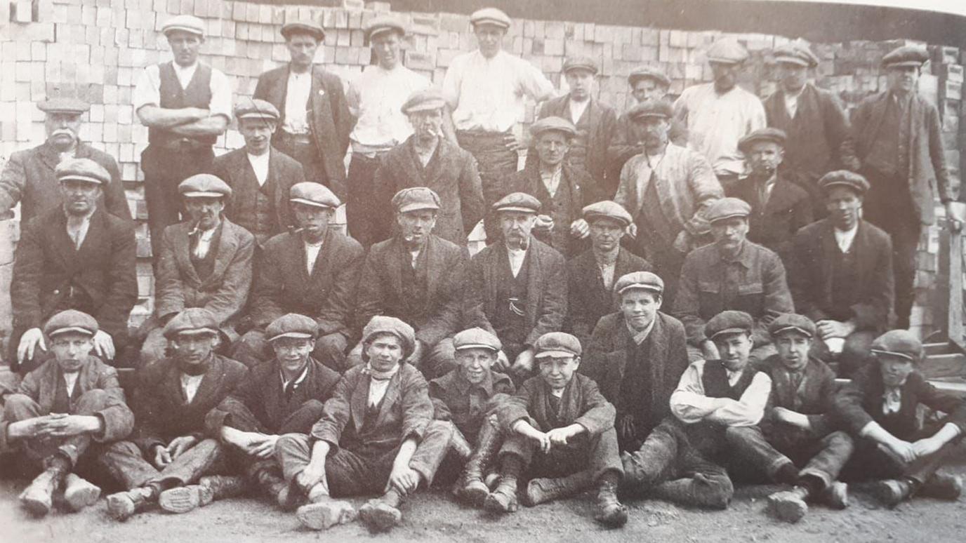 Black and white photo of boys and men lined up in four rows at Dogsthorpe brickworks, Peterborough, in about 1900, all wearing flat caps