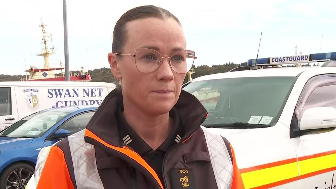 Roisin McBride is standing outside speaking to the media. She wears glasses and has a coastguard uniform on. A boat and cars are behind her. The sky is cloudy.