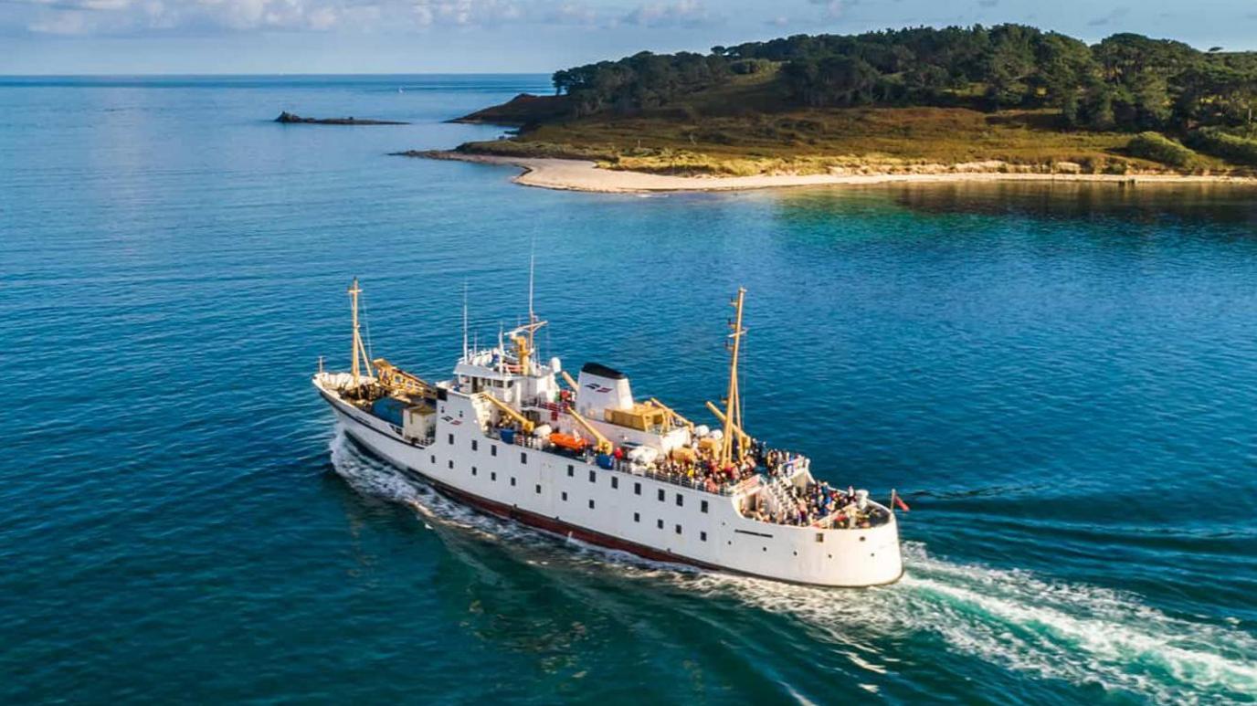 The Scillonian ferry, with passengers on board, from above, going past the edge of an island