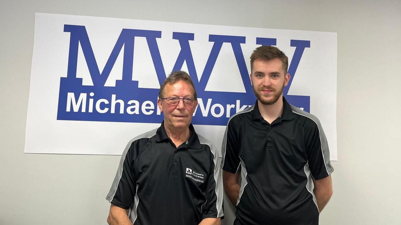 Michael Titcombe and Luke Webster in work uniform standing in front of a Michael's Workwear sign