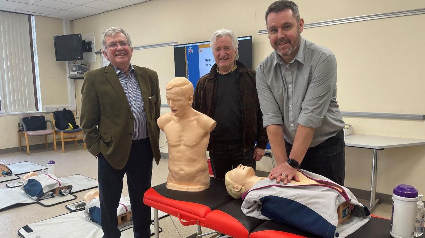 Three men standing in a hospital behind two medical mannequins