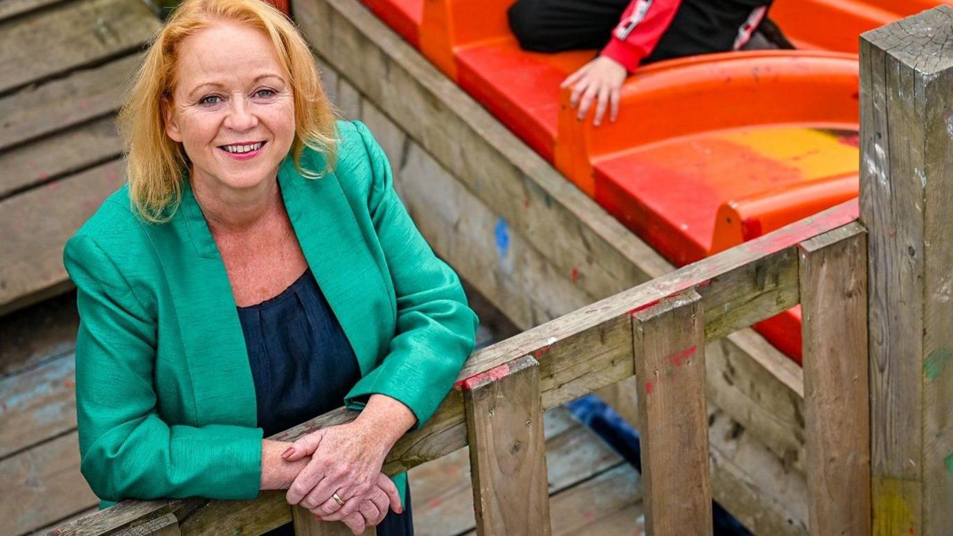 A woman smiling and looking at the camera. She is wearing a green jacket and leaning on a wooden fence. A child is on a slide in the background. 