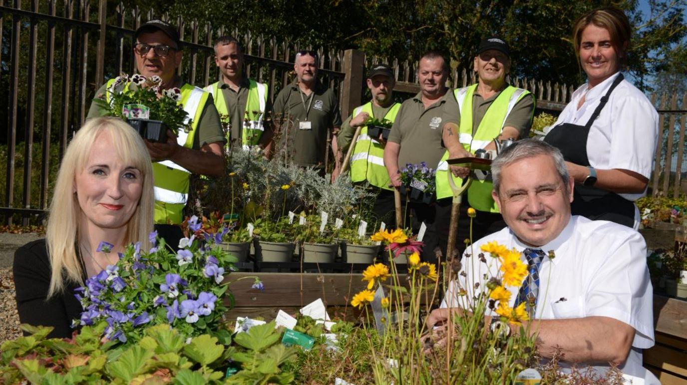 Kelly Chequer and Shaun Donnelly crouch behind a planter filled with flowers. Volunteers stand in the background holding flowers.