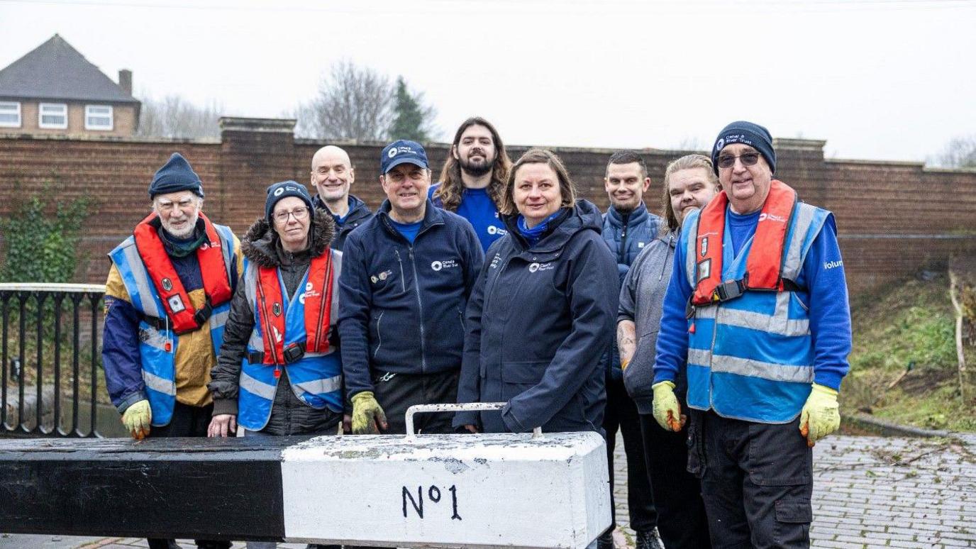 Nine men and women in stand in front of a canal lock, smiling at the camera. They are all wearing blue Canal and River Trust uniforms and some have orange inflatable safety jackets on.