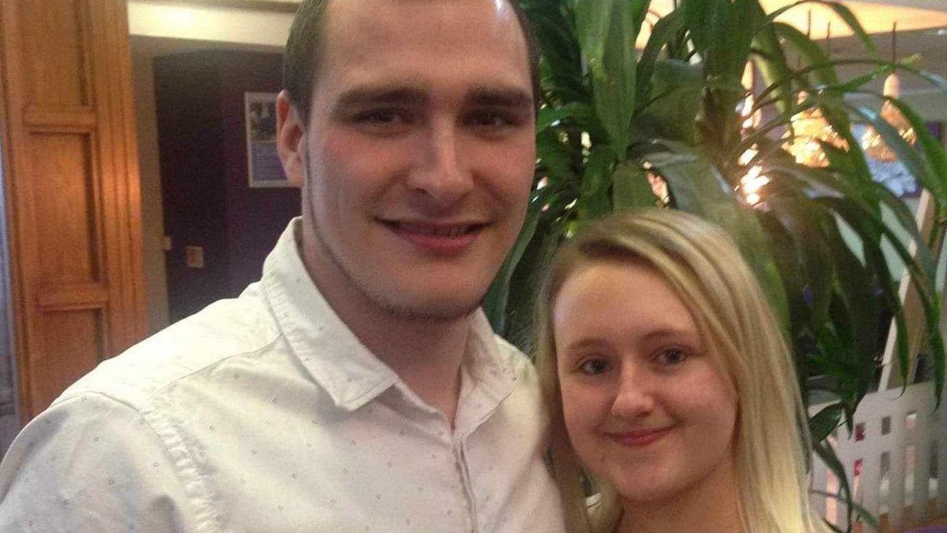 Smiling Chris Taylor, who has brown hair and a beard and is wearing a white shirt, is pictured with his partner Jemma, who has long blonde hair and minimal make-up, in the foyer of a venue