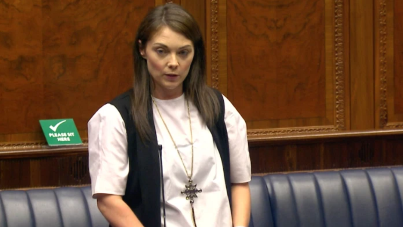 Jemma Dolan speaking in the Northern Ireland Assembly. Behind her the blue benches and a wood-panelled wall. She is wearing a white shirt, black waistcoat and large necklace 