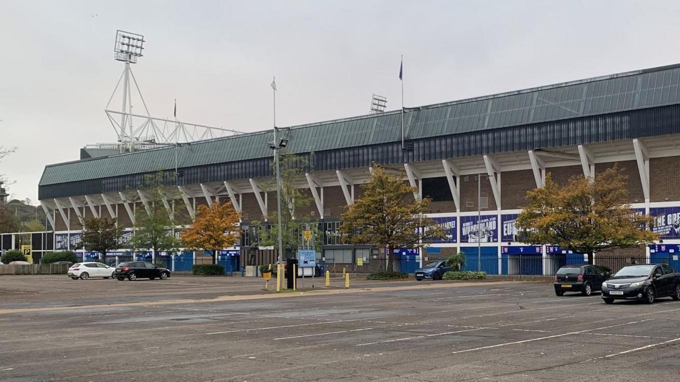 Portman Road, as seen from New Portman Road car park. The two-tier stadium can be seen in the background, with its black roof and white supporting columns outside. In the foreground are cars parked in a car park on a cloudy day.