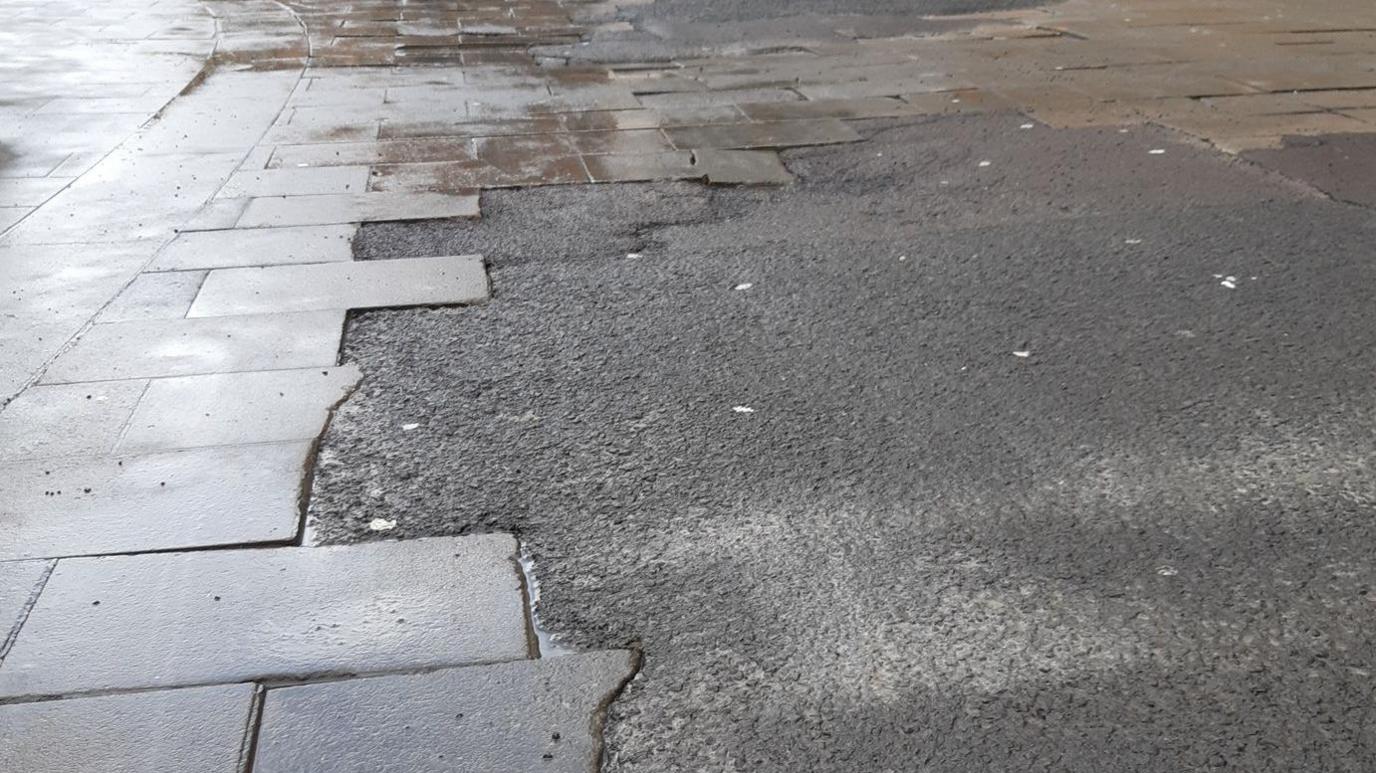 A still image of Queens Street in Oxford. There are paving slabs that are disjointed and there is tarmac scrambled around them.