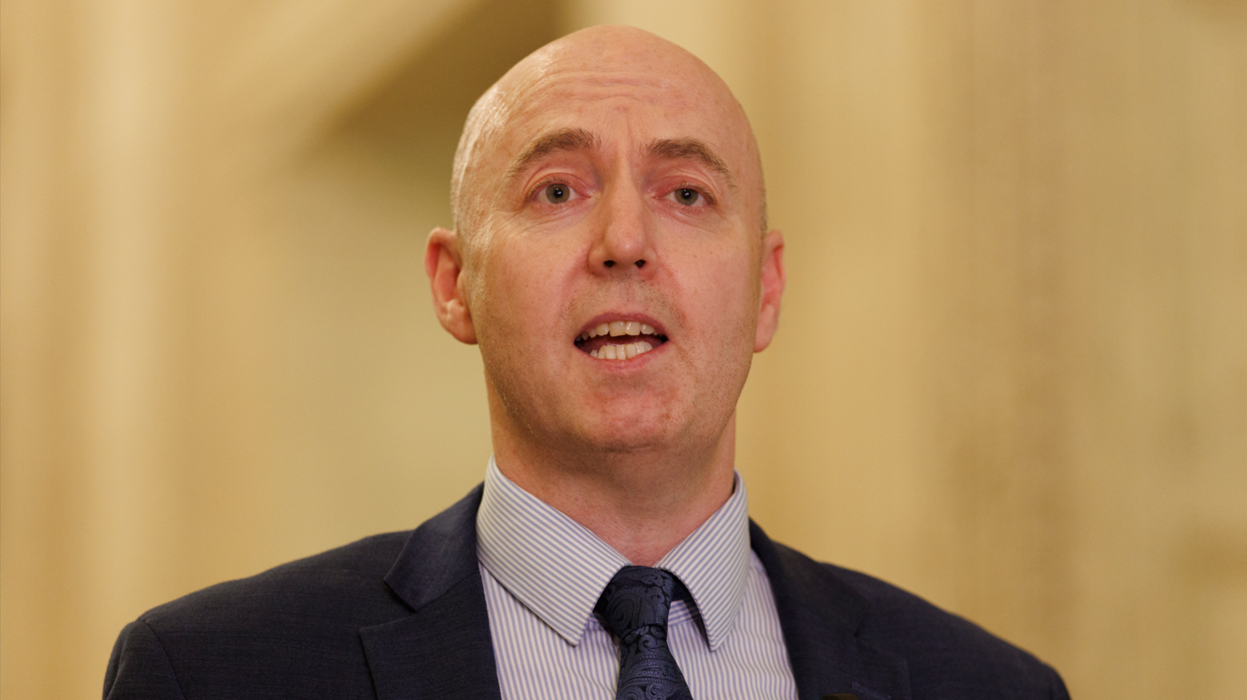 Jon Burrows is talking. He's wearing a navy suit with a white and blue stripped shirt and a navy tie with a pattern. The background is out of focus.