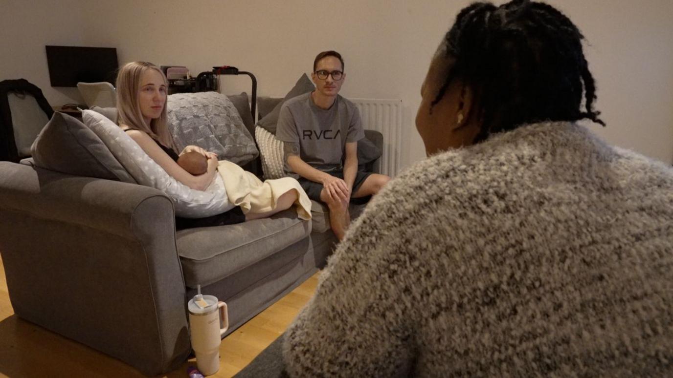 Mosun isviewed from behind seated and speaking with a family in their living room. A mother holds a baby wrapped in a blanket on a grey sofa, while a father sits beside her.