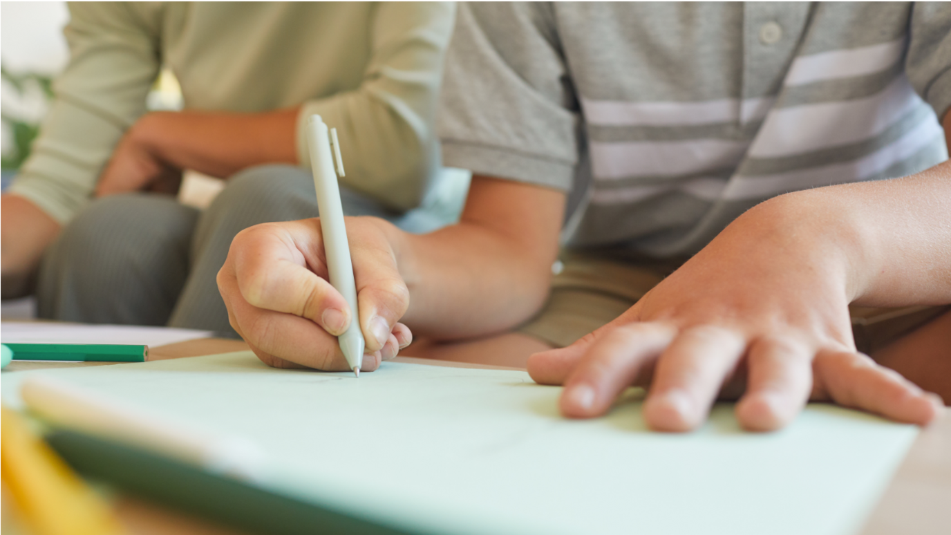 An unidentified child writes on a board with a pen. An adult sits close behind them.