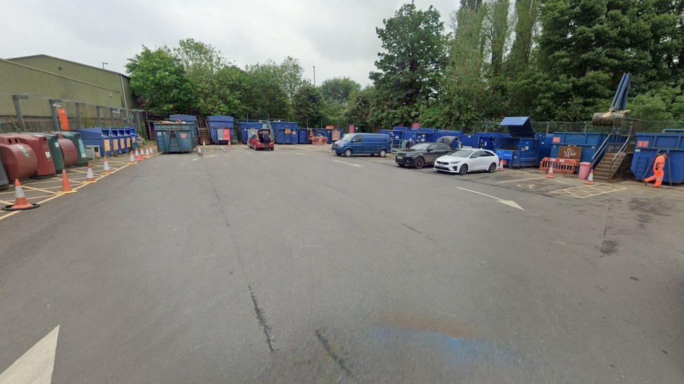 A car park at a waste recycling centre, with bottle banks on the left side and waste containers on the right. Cars are parked up, with domestic waste being carried to the blue skips. 