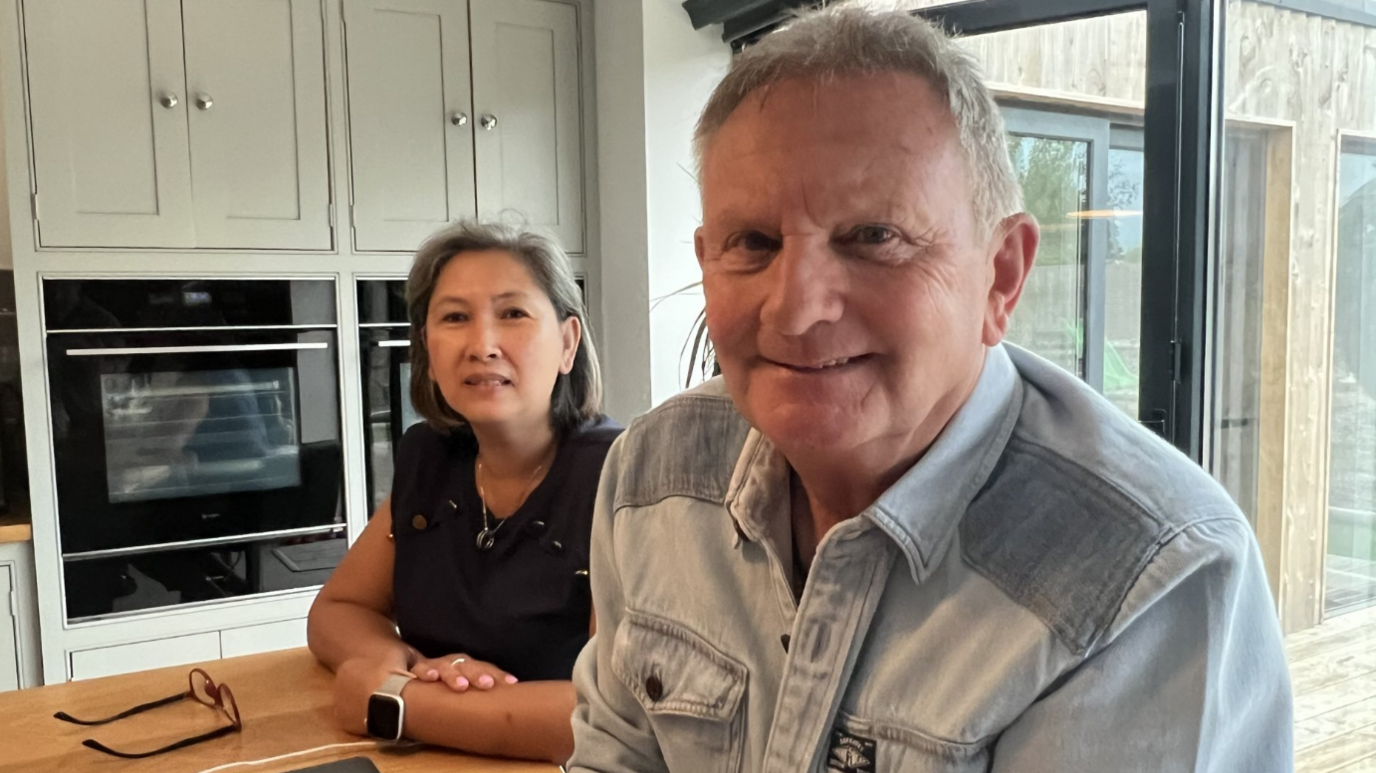 Kev and Mae Draper smile at the camera as they stand together in their kitchen