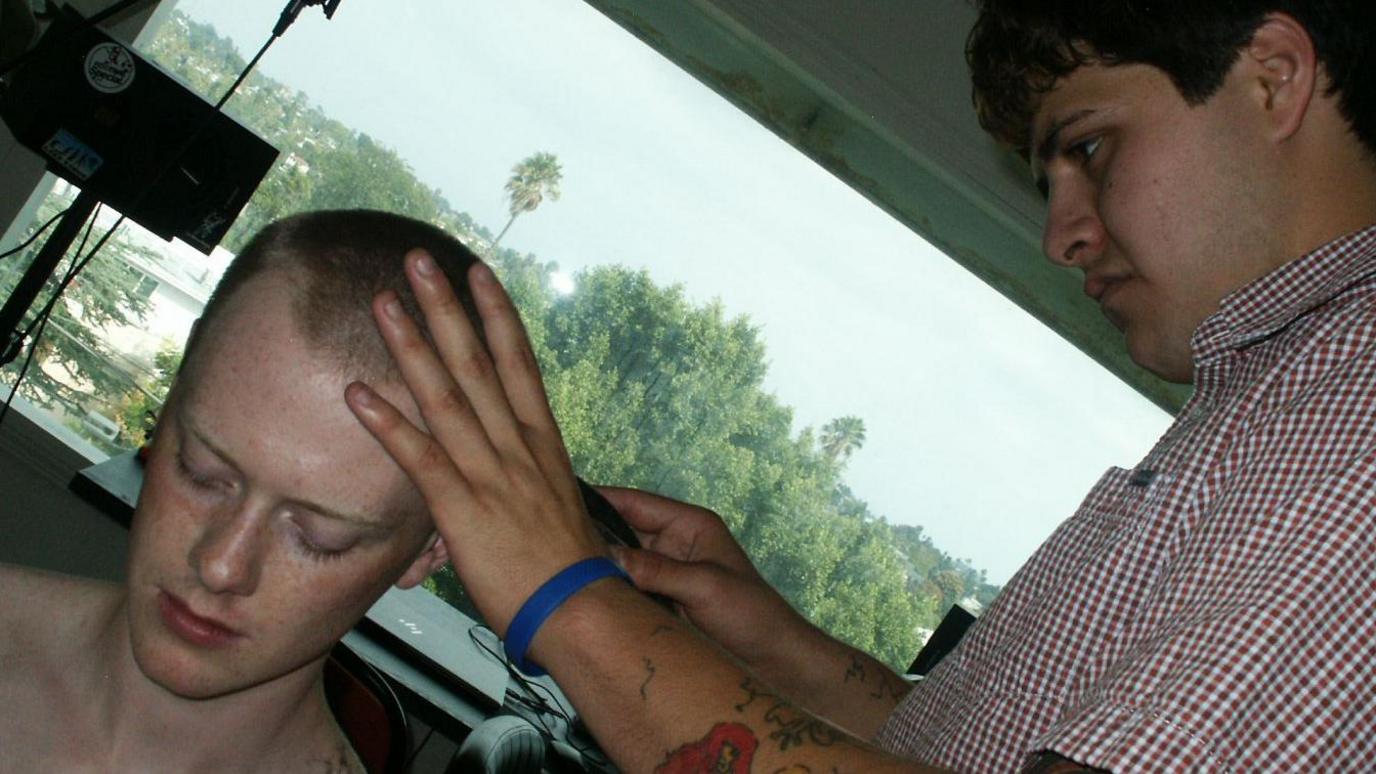 A man sits as he gets a haircut. He is in the bottom left corner of the picture which is tilted. His hair has been shaved to a buzz cut. Another man wearing a patterned shirt is cutting his hair. That man has brown hair and is holding the razor in one hand with his other positioned on the first man's head. A window can be seen in the background revealing a bright day in LA full of trees.