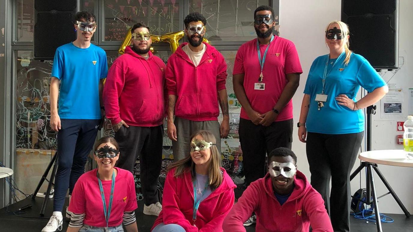 Three people wearing pink hoodies and T-shirts stand next to two wearing blue T-shirts, with another three in pink sat in front of them. They are all wearing silver, gold or black masquerade-style masks.