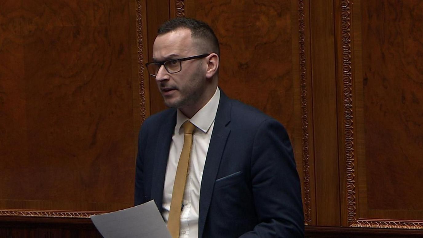 Timothy Gaston speaking in the Stormont Assembly. He has dark hair, wearing glasses, a blue suit, white shirt and yellow tie.
