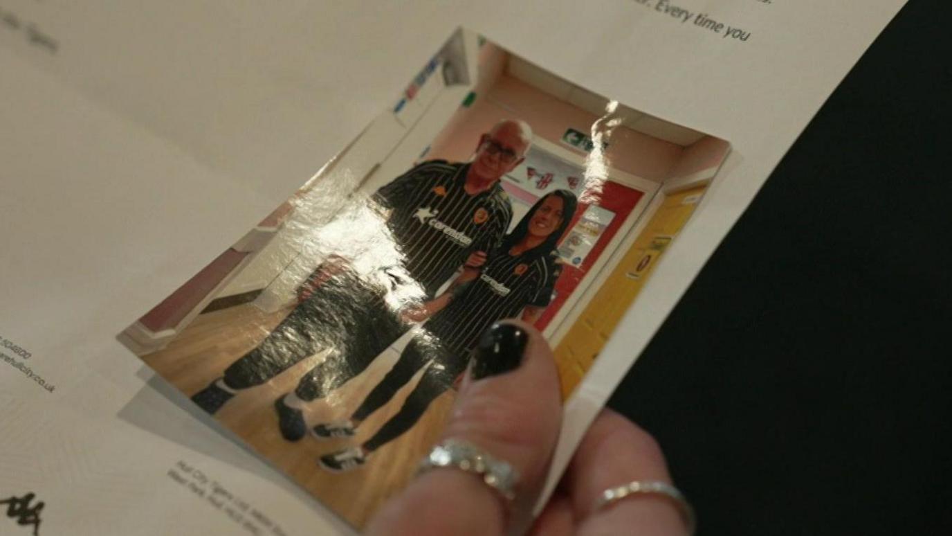 A hand with silver rings and nails painted black holding a photo of Sophie Griffiths and her father. They are both wearing Hull City FC kit and are stood in a corridor. Sophie's dad has a bald head and is wearing black glasses.