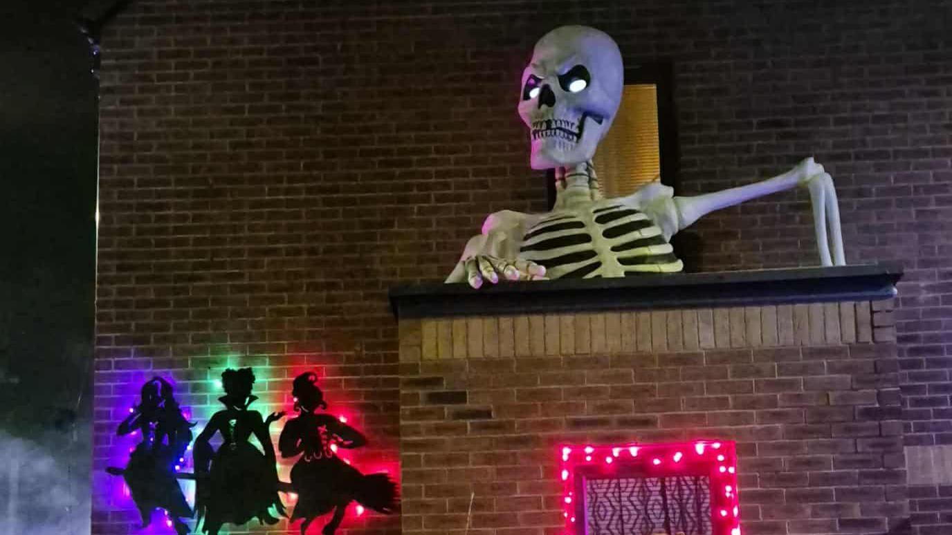 A silhouette of the Sanderson sisters from Disney's Hocus Pocus on the side of a house set against purple, green and pink lighting. There is also a giant skeleton appearing above a flat roof on the side of the building and a window illuminated with pink lights around it.