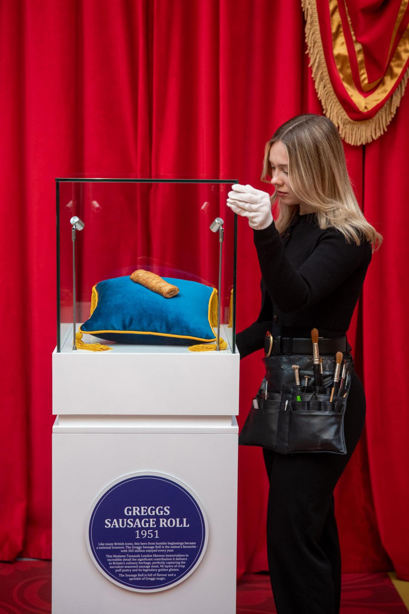 A woman fixes the sausage roll into a glass case on a plinth 