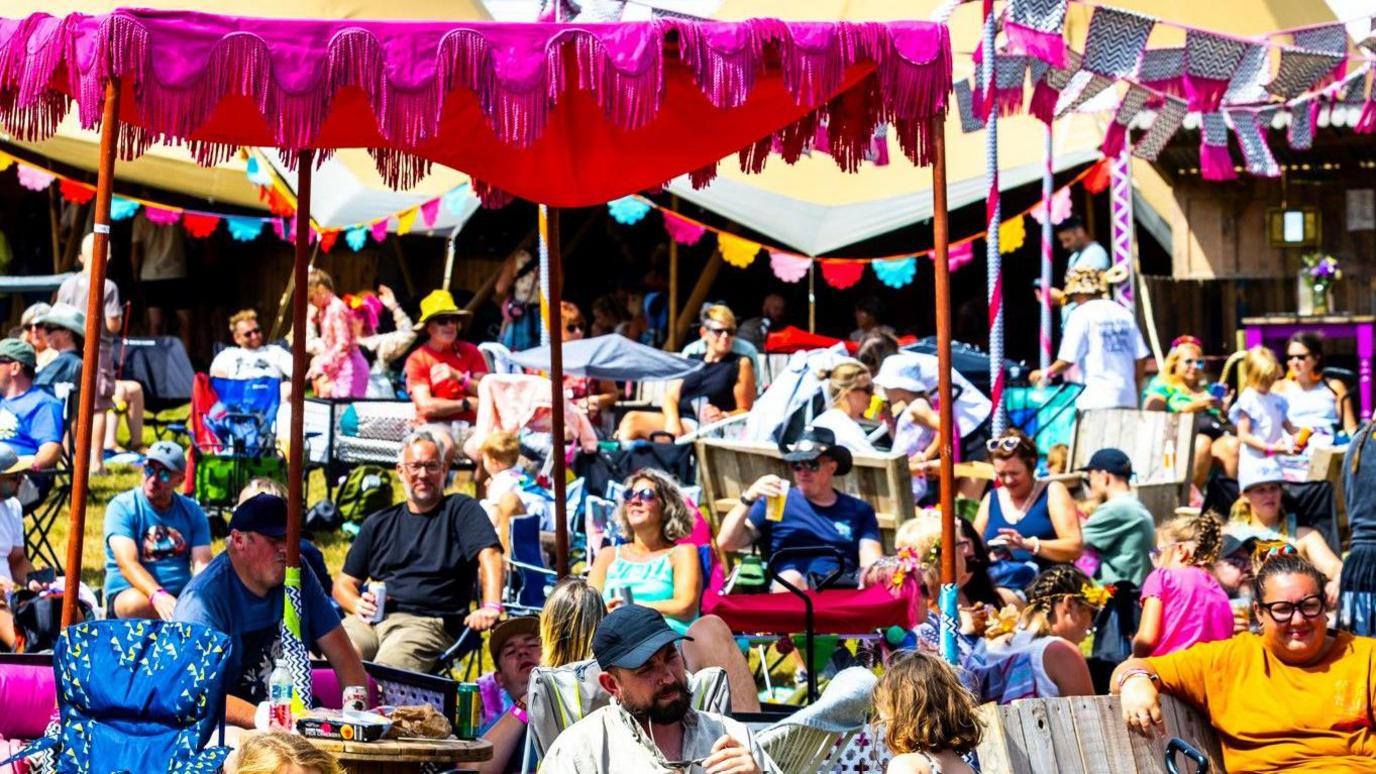 A telephoto shot of lots of people sitting in the sunshine in front of some brightly coloured tents decorated with bunting