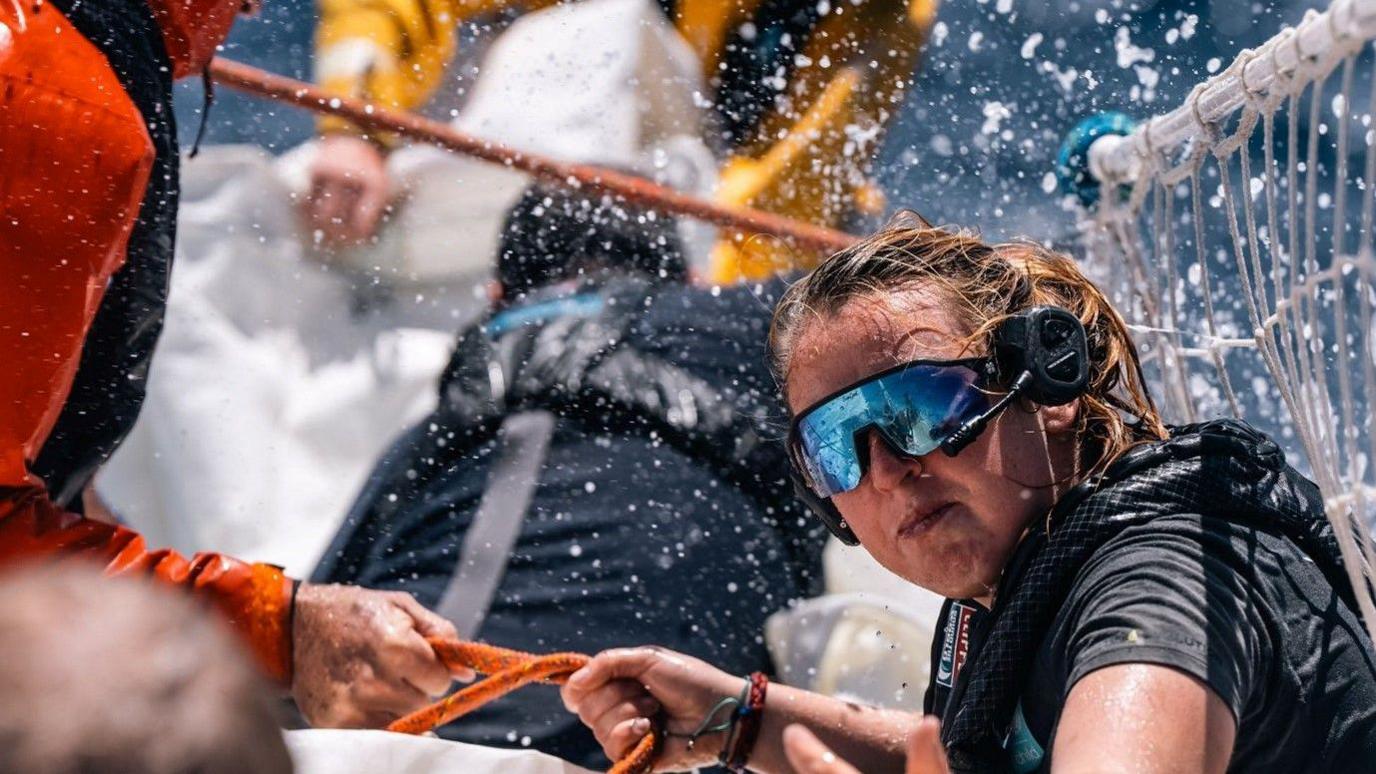 Image from last year's race, of a young woman in action on board a yacht in wet gear and glasses battling ocean spray.
