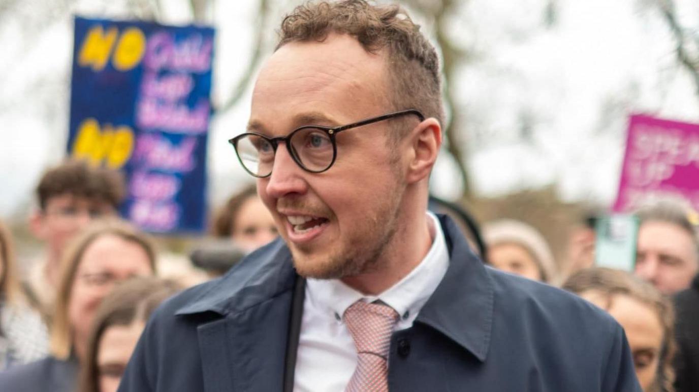 Adam Dance is standing in a crowd of people. He is the only one in focus. He is wearing a navy jacket over a suit with a pink tie. He is saying something to someone behind the camera.