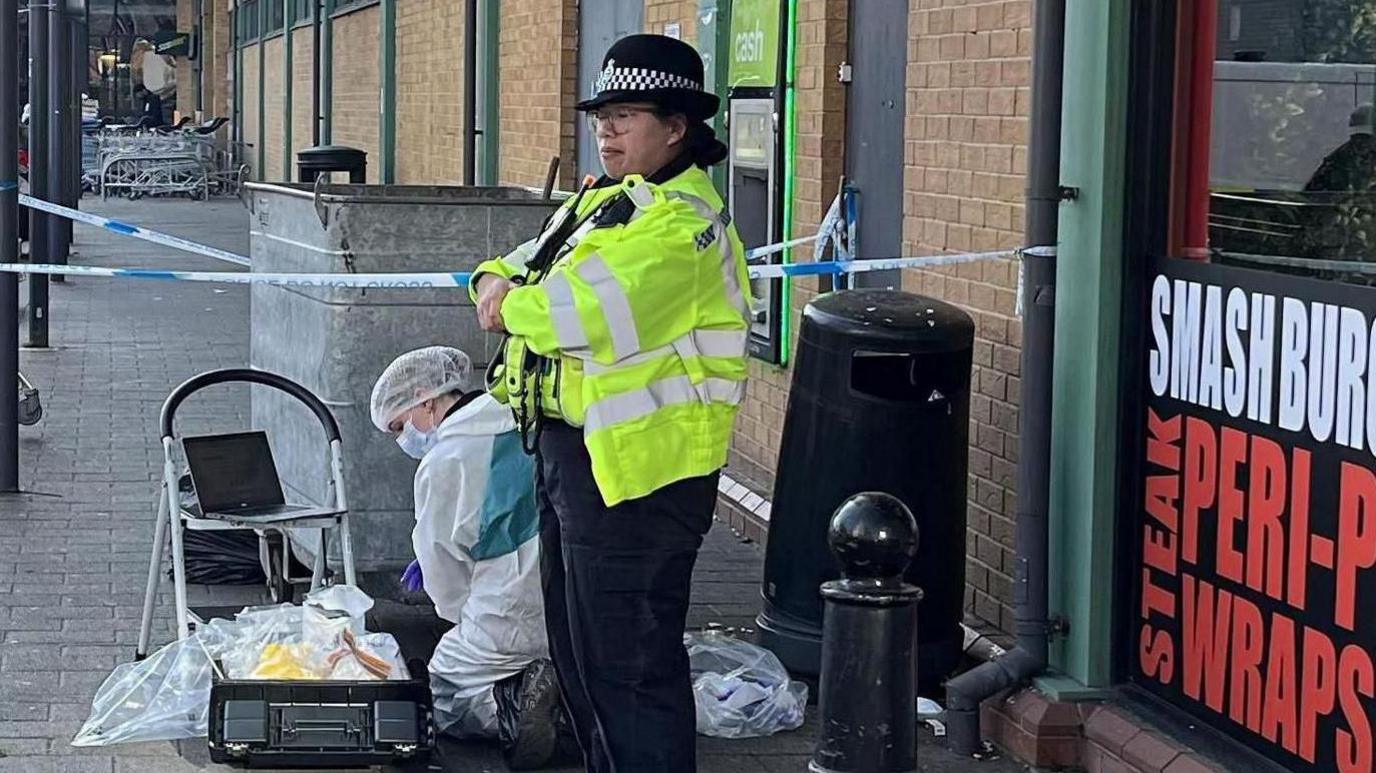 A woman police officer wearing a yellow coat stands with folded arms outside a fast food shop. Kneeling to her right is a crime scene investigator dressed in a white suit.