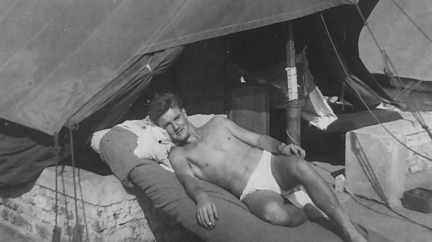 Gerry Bowler, wearing only pants, lounges outside a tent, in a black-and-white photograph from the 1950s.