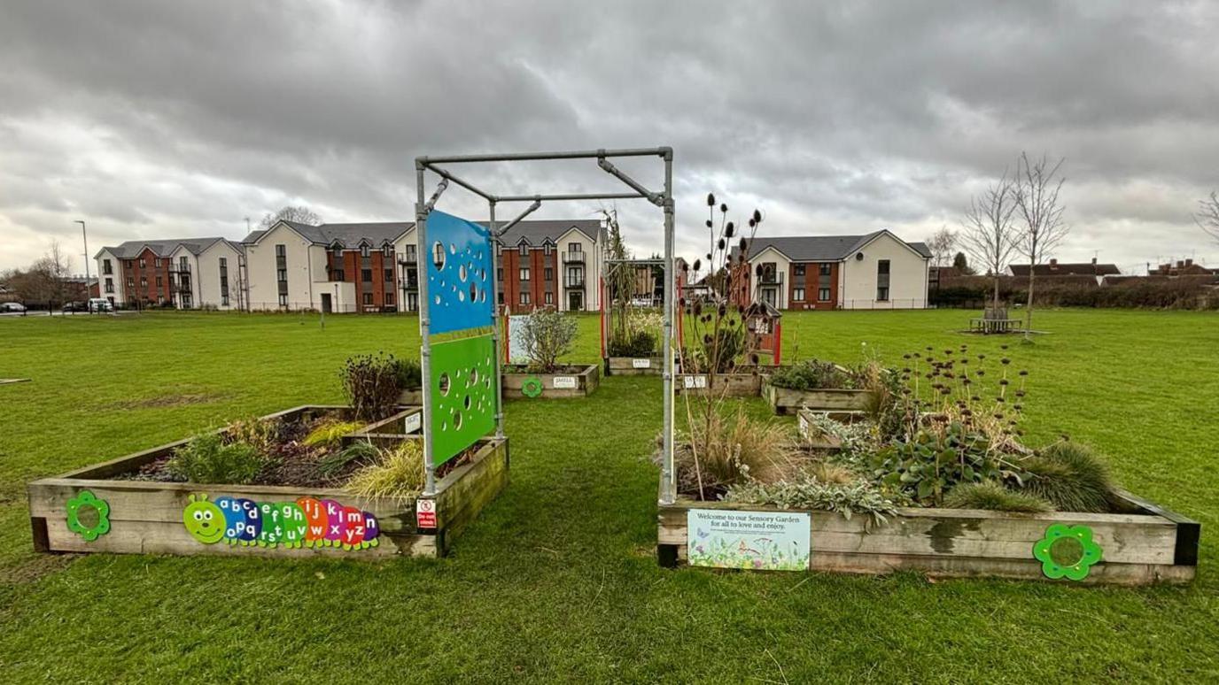 The sensory garden, set in a large green space with flats visible in the background. There are a number of large raised planters and a metal archway. 