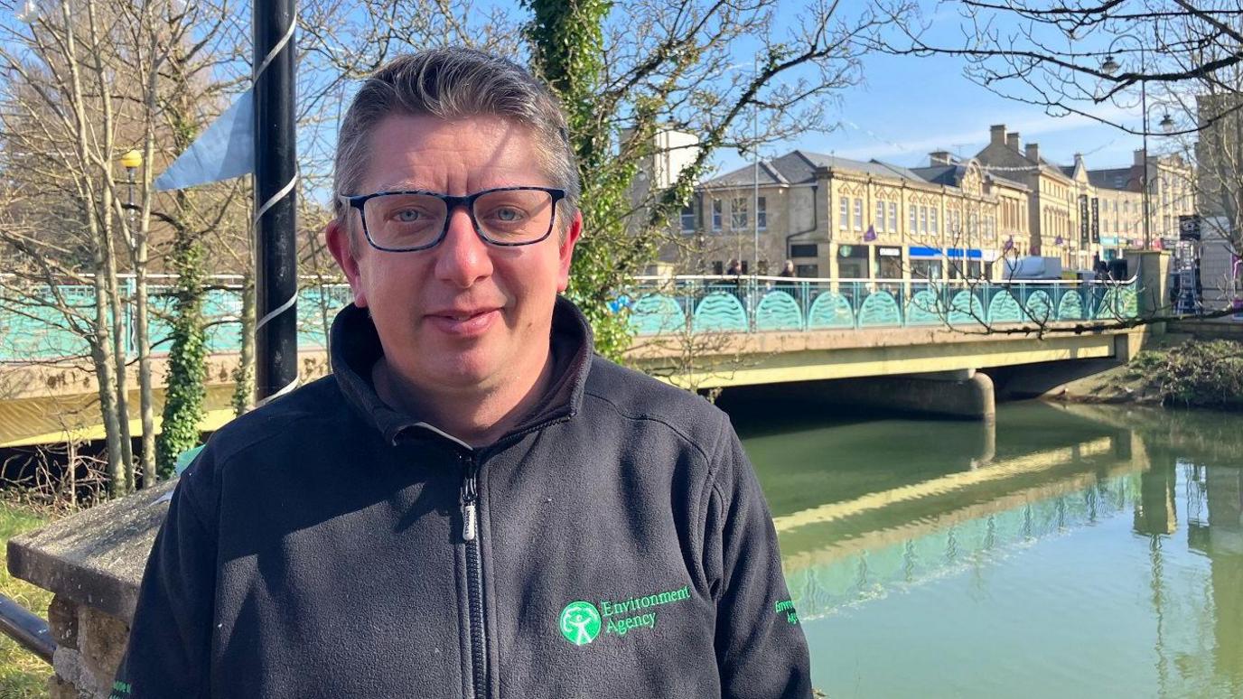 Andy Wallis - wearing glasses and an Environment Agency branded fleece - looks at the camera while standing in front of a river and bridge.