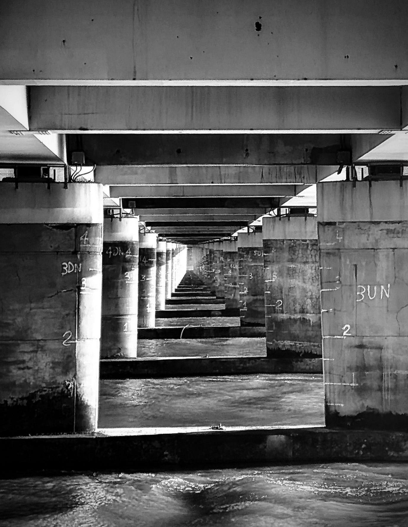 A black and white scene underneath a bridge with pillars and water lapping below 