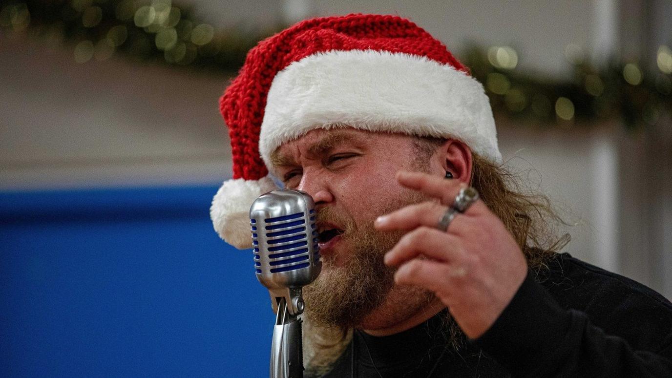 Callum Doignie has long brown hair and brown facial hair. He is singing into a microphone with his hand by his face. He is wearing a red and white knitted Santa hat