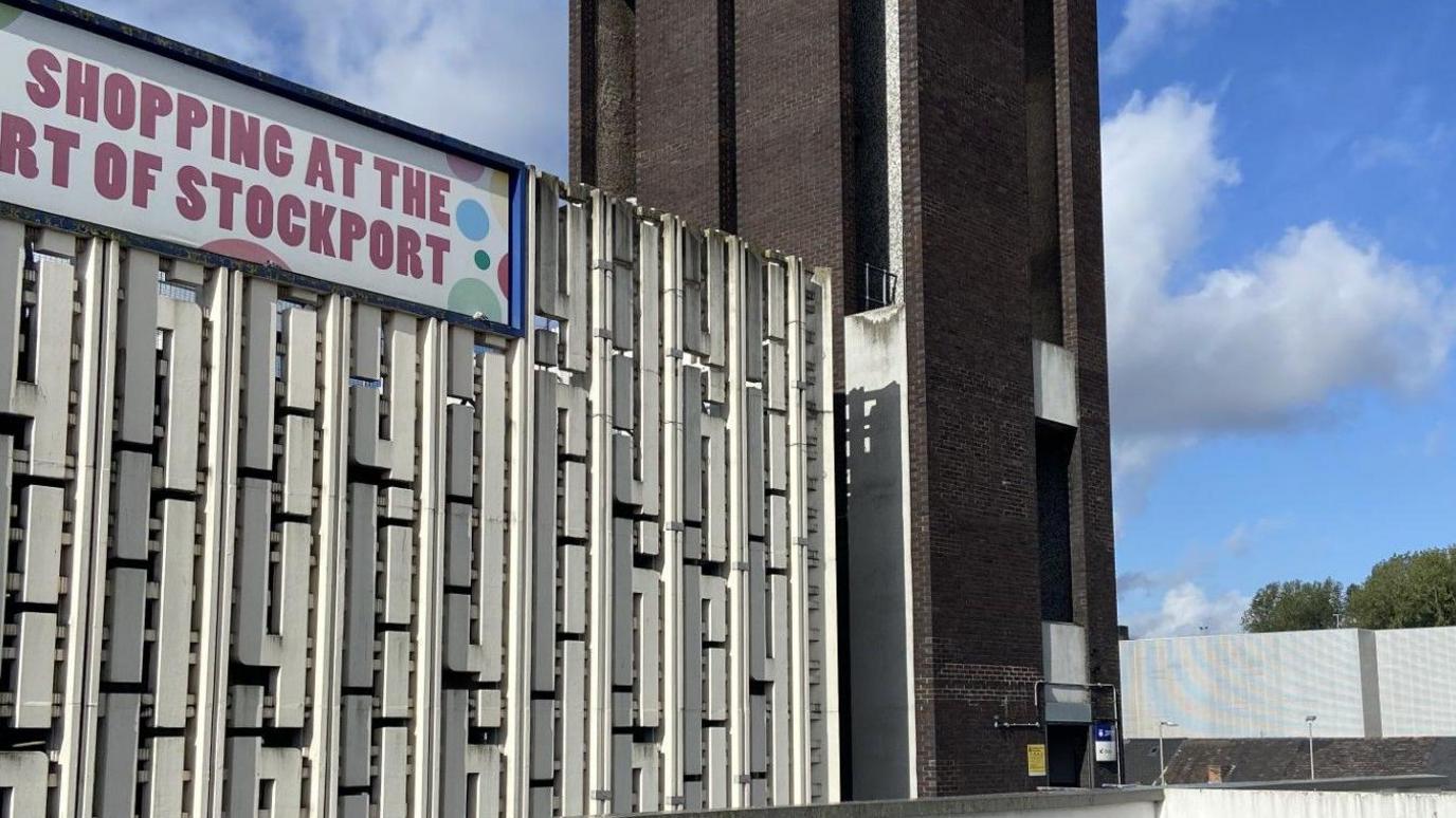 A light grey rectagular building with thin cutout sections. It is next to a brick lift shaft and has a banner hanging over the top that reads Merseyway: Shopping at the heart of Stockport" but on the picture the writing is cut off 