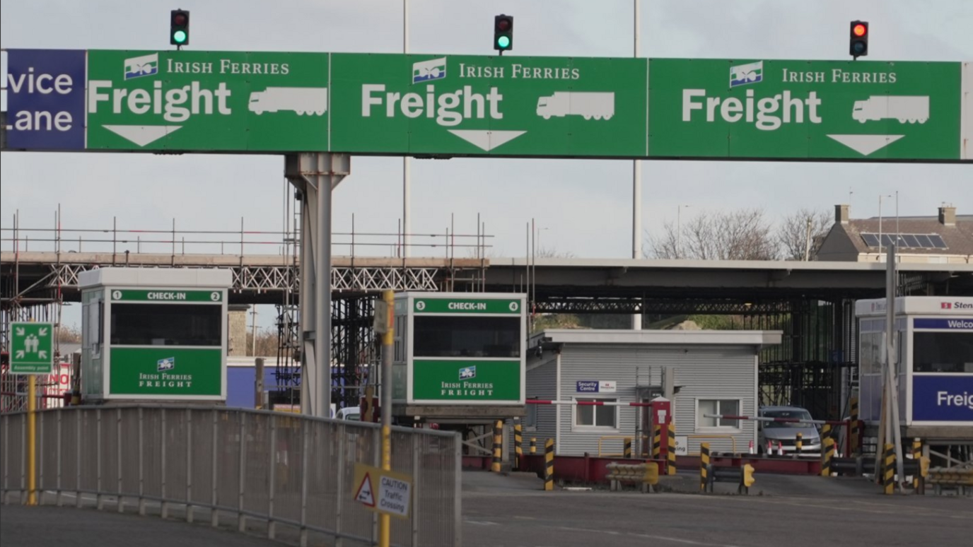 A view of Holyhead Port in North Wales. We can see the overhead green road signs for motorists to know which lane to enter. Labelled 'freight'. We can see checkpoint boxes with the word 'check-in' above. Lifting barriers separating the two sides of the roads.