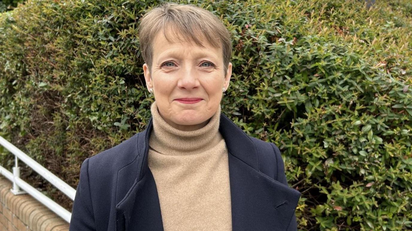 Clare Moody looks at the camera. She has short dark hair and wears a fawn turtleneck jumper and a navy overcoat. She is standing in front of a large bush. 