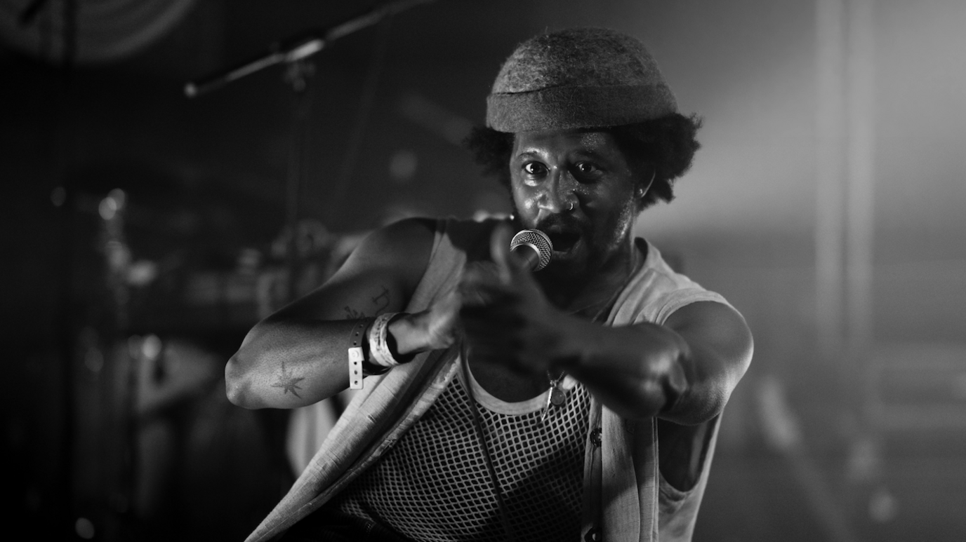 Artist Hang Linton wears a woolly hat, string vest and sleeveless top. He is singing into a microphone
