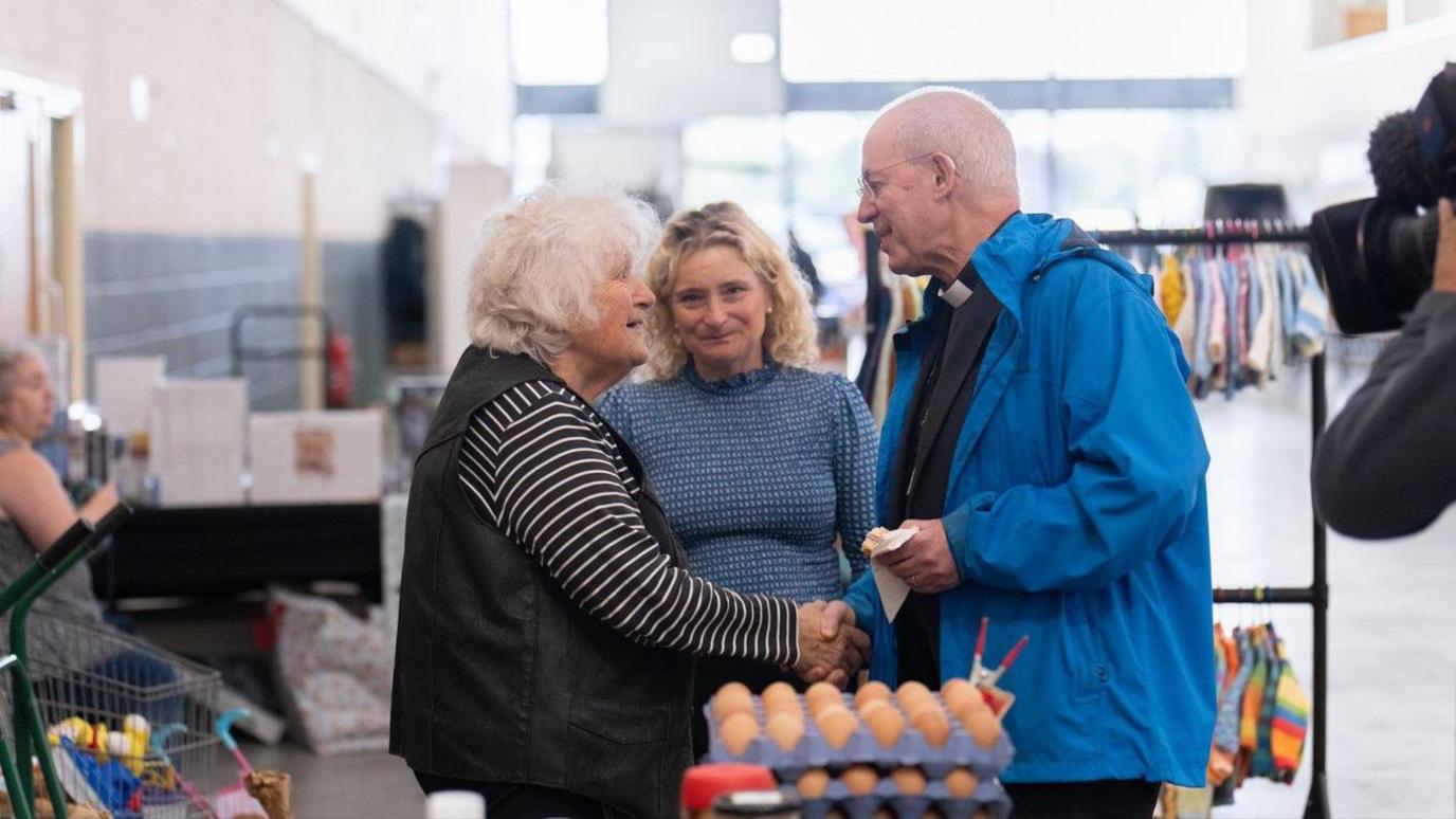 The archbishop (right) talking with Ms Adams (left) at the market. He is wearing a blue coat and there is food produce and other items in the foreground.