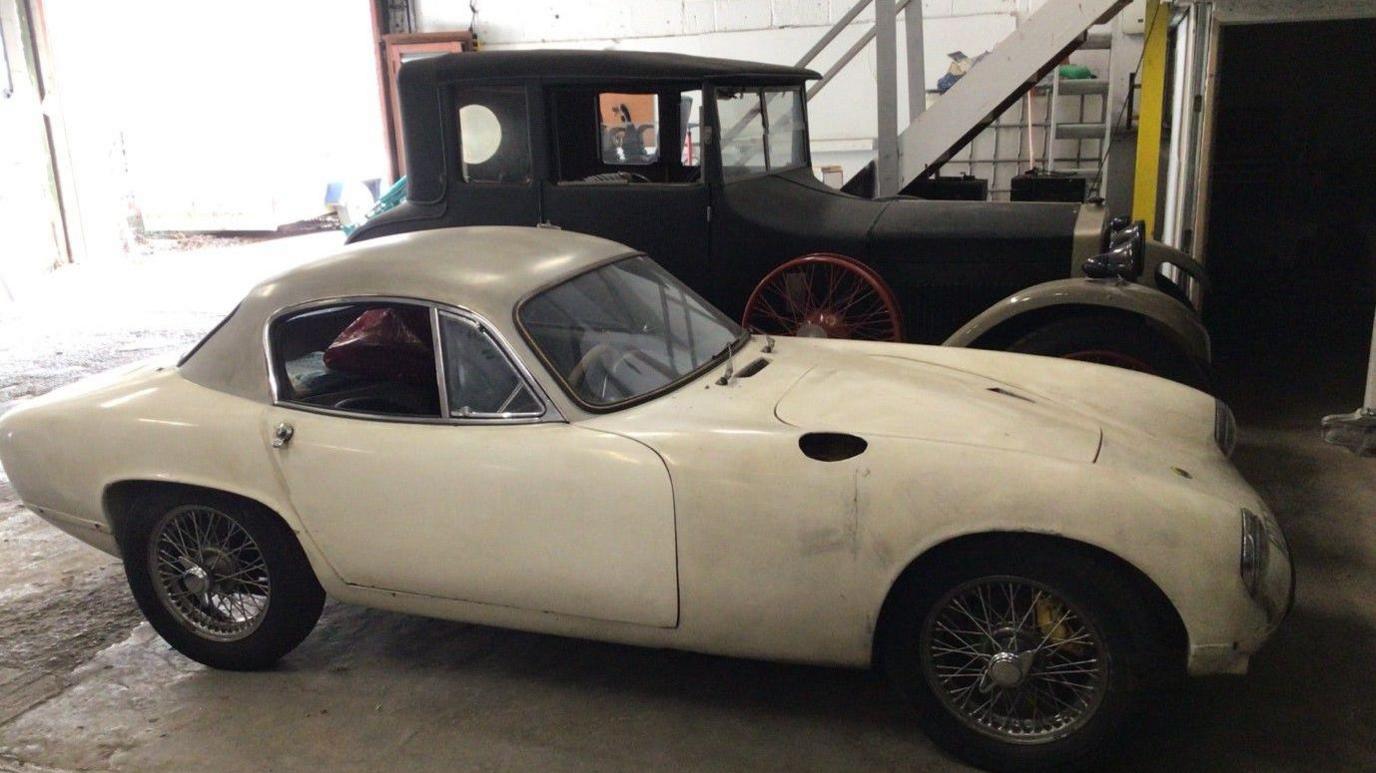 A white two-door Lotus with a tan roof. It is parked in a barn next to the black Diatto.