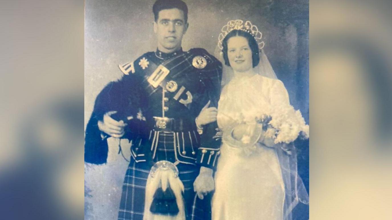 Private Robert Balfour, a dark-haired man, with his wife Doris, who also has dark hair. He is wearing a Scottish soldier's ceremonial uniform, including a kilt. She is wearing a long, white wedding dress, with veil, and is carrying a bouquet of flowers. They are arm in arm. 