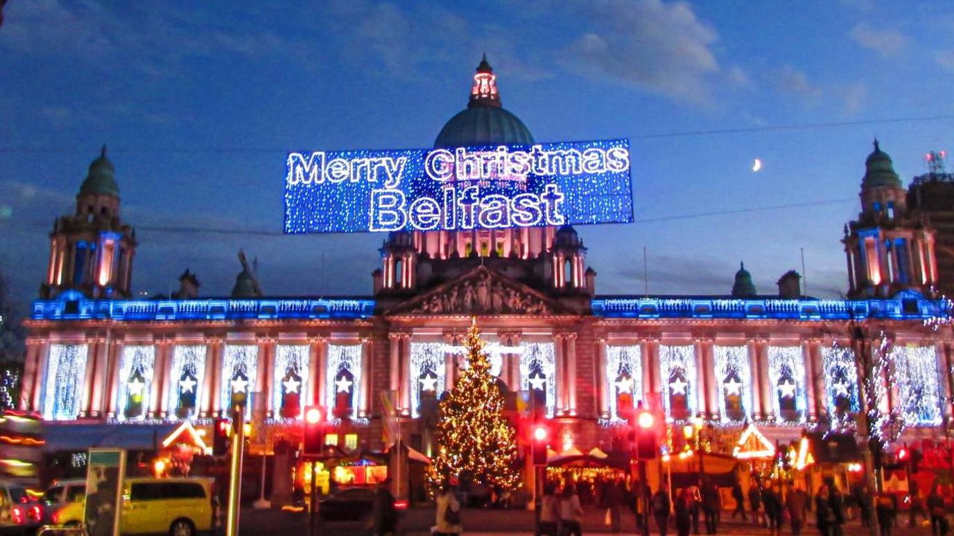 The exterior of Belfast City Hall with it's Christmas lights on, as well as some wooden chalets welcoming visitors as part of its Christmas market. There is a large lit-up sign saying "Merry Christmas Belfast"