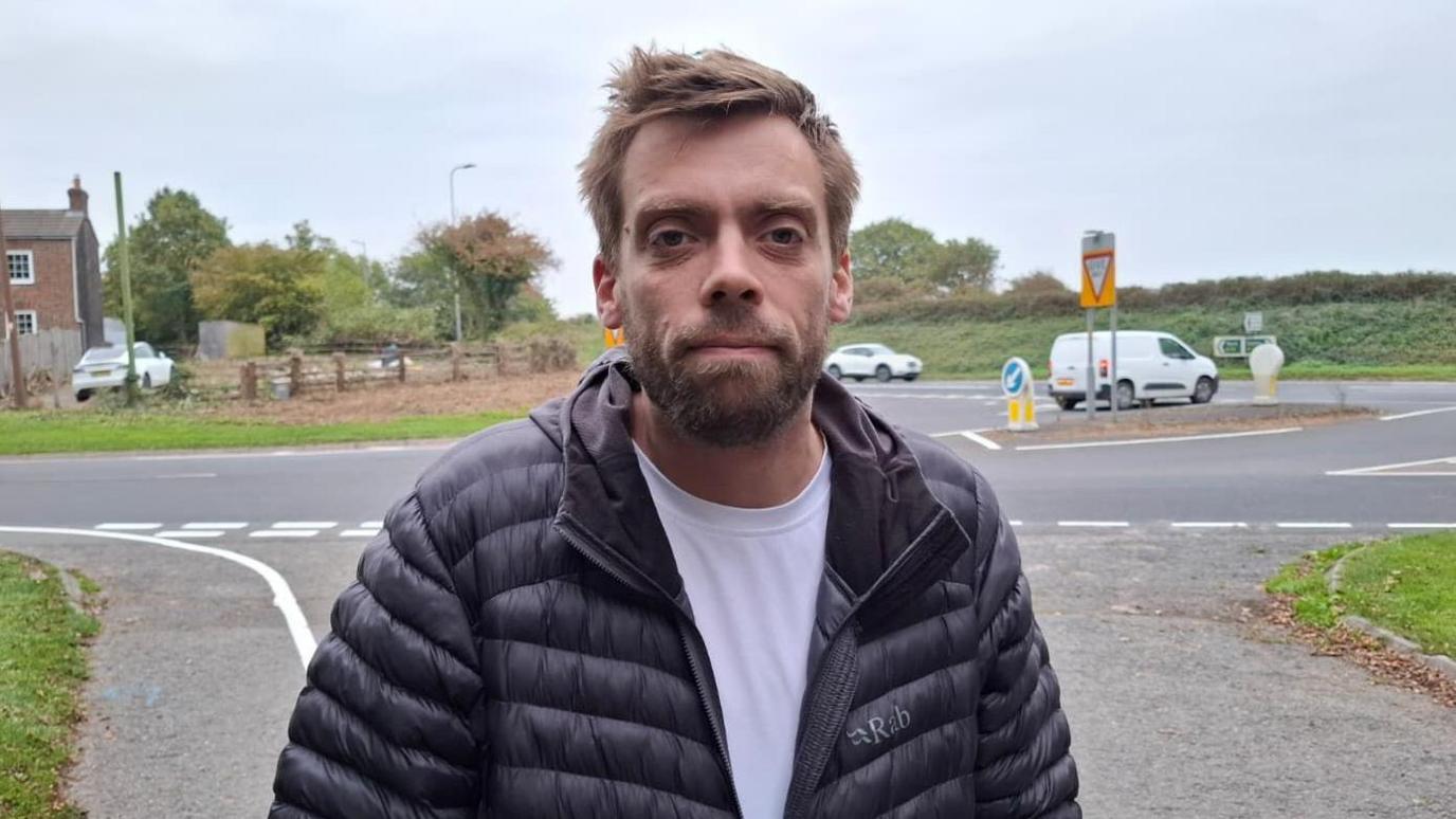 A man with brown hair and a beard looks at the camera with a neutral expression on his face. He is wearing a black jacket and white T-shirt. In the background is a busy road junction leading onto the A19.