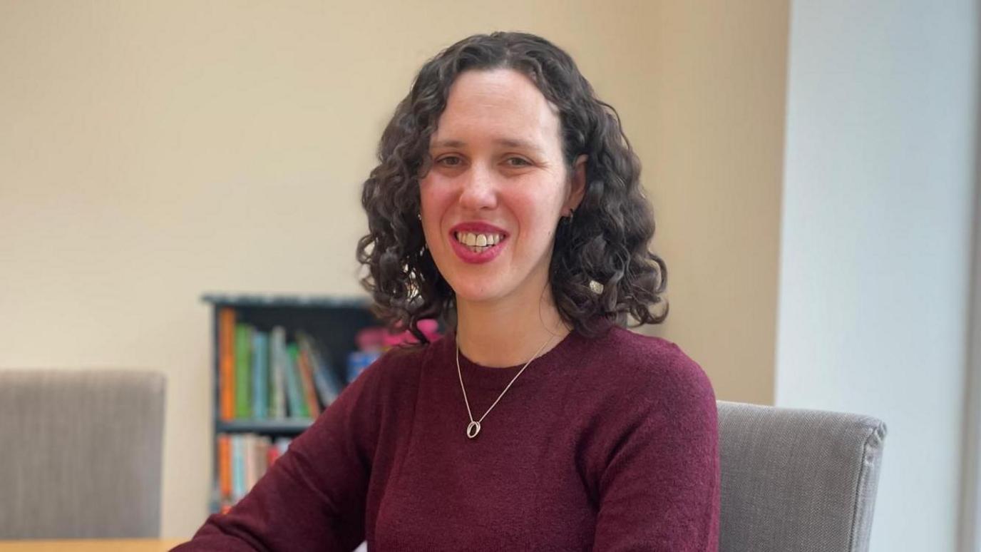 Mae Georgina Forbes gyda gwallt cyrliog du i'w ysgwyddau ac yn gwiso ffroc coch tywyll. Mae hi'n gwenu ac yn eistedd wrth bwrdd cegin gyda silff llyfrau yn y cefndir.