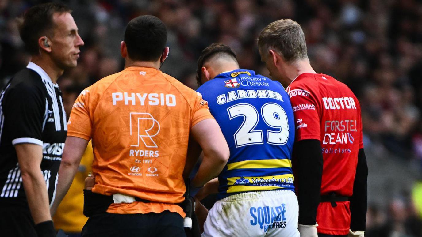 Zak Gardner (GARDNER 29) is helped from the field by a PHYSIO (orange top, left) and a DOCTOR (red top, right) during Warrington's win at St Helens