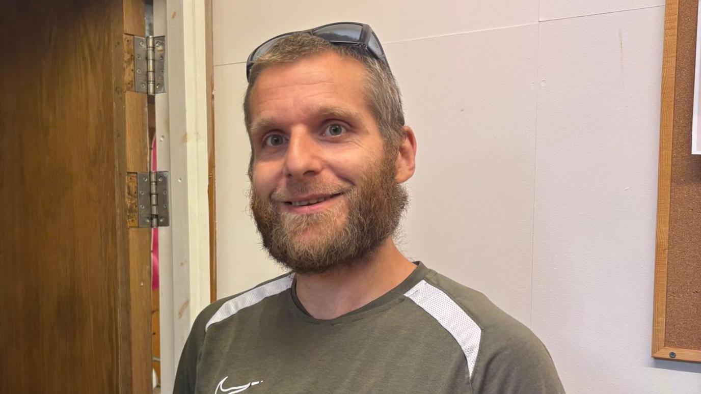 A white man with brown hair and a beard. He is wearing a green striped t-shirt with sunglasses on his head.