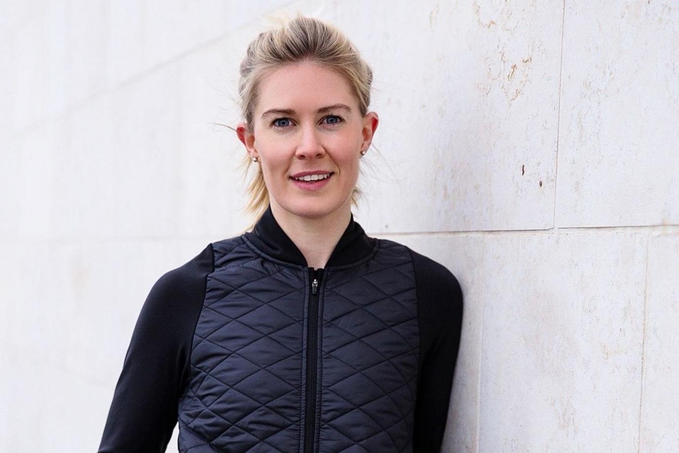 A woman with blonde hair smiles as she leans against a white wall while wearing a black gilet.