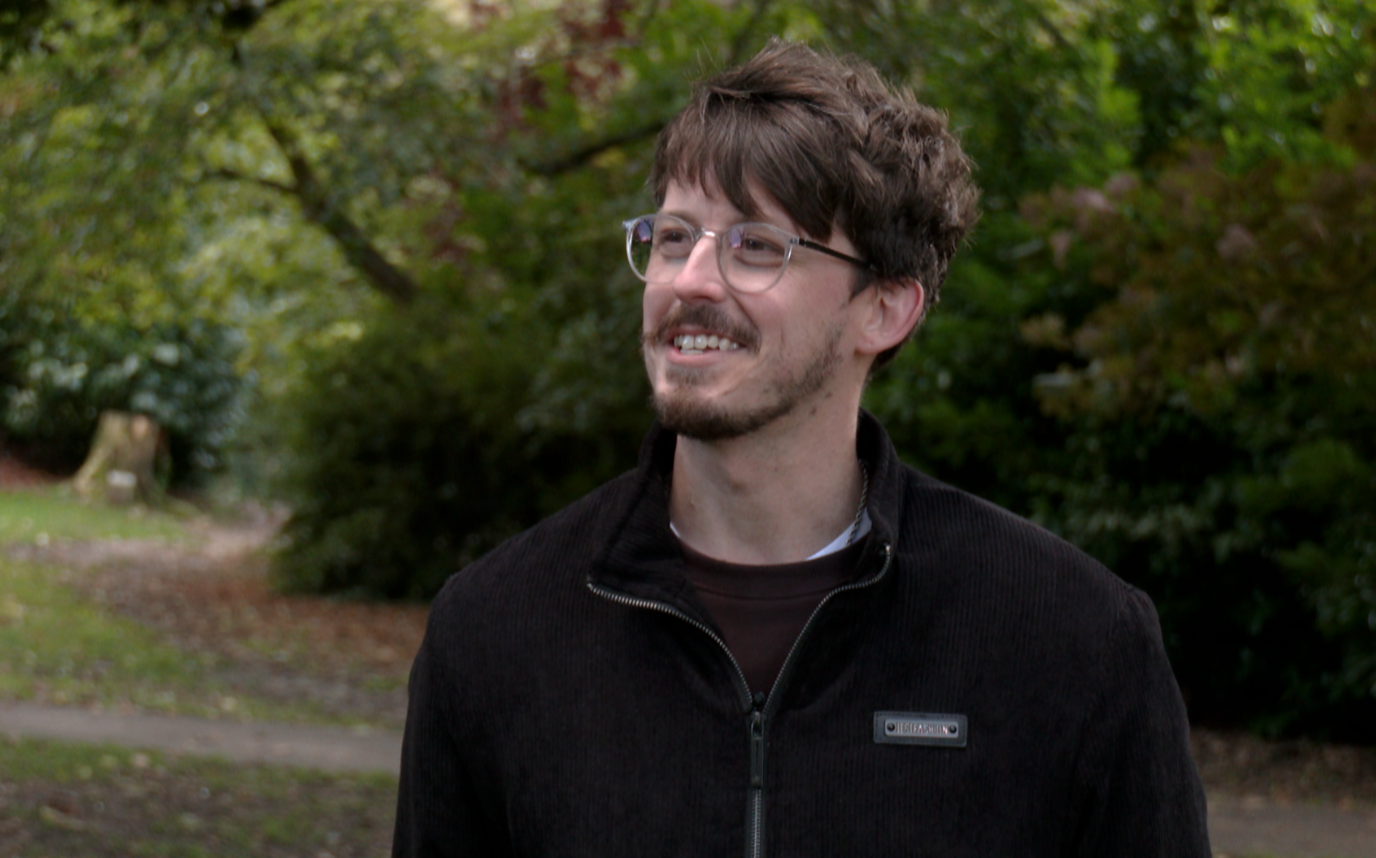 Luke Cowdell wearing a black zip up top in a park, surrounded by bushes and trees