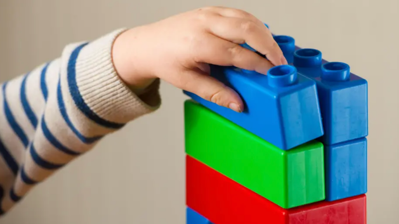 A child's hand stacking large building blocks. The blocks are blue green and red.