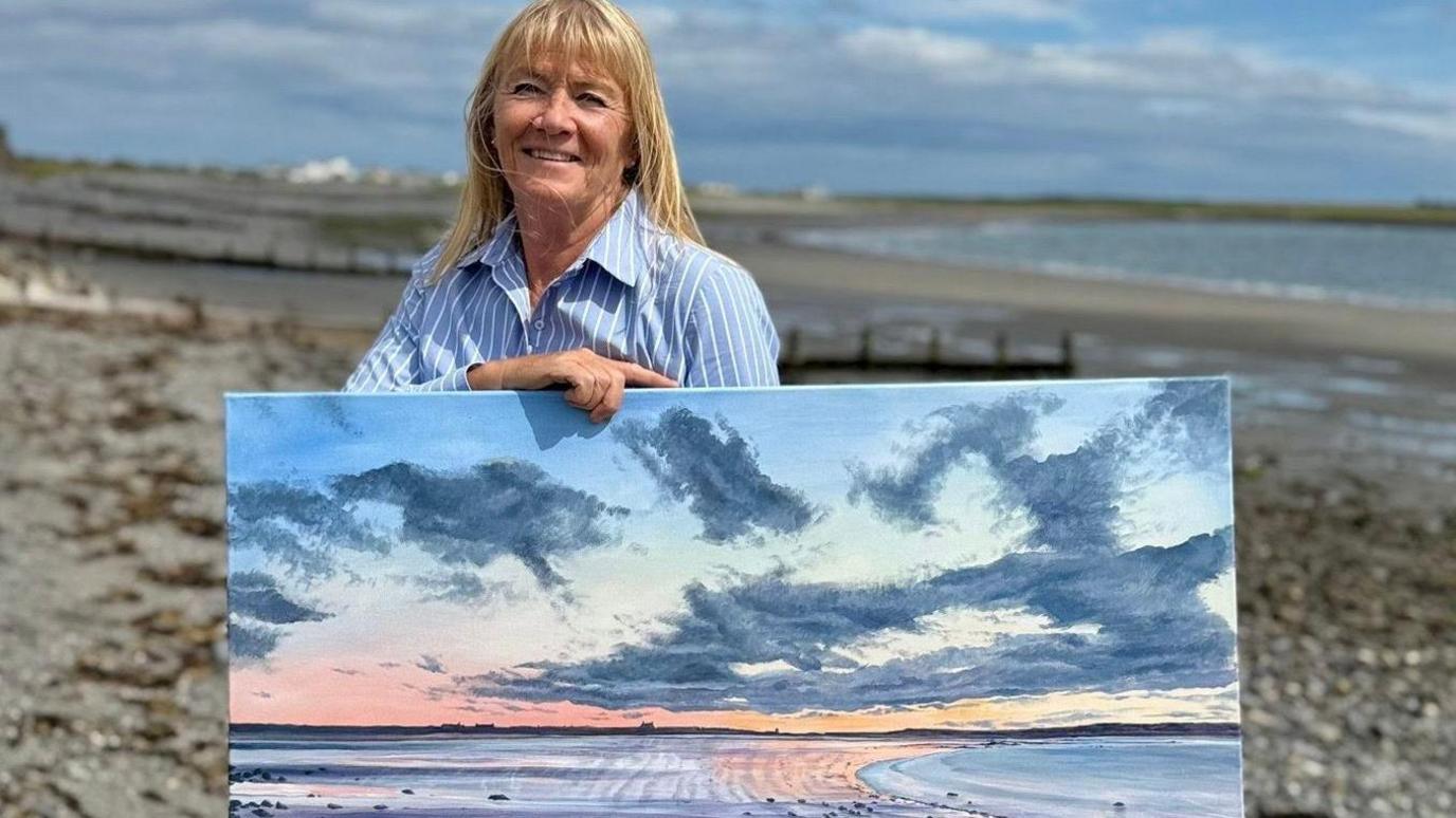Chrissie Moss, a woman with blonde hair and a fringe, she smiles and wears a blue stripy shirt, as she stands on a wide beach, holding a painting of that same beach at sunset.